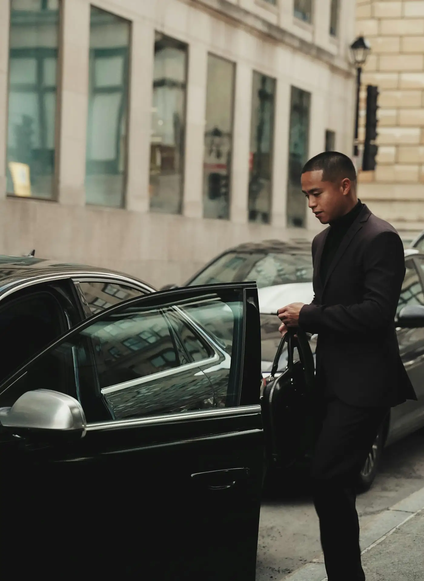 Man in a dark suit opening the rear door of a black car on a city street