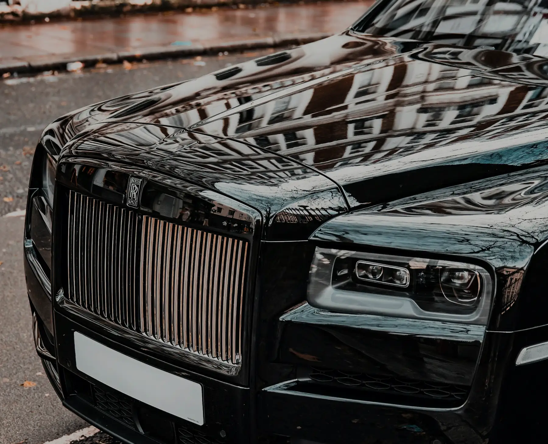 Front of a black Rolls-Royce Cullinan on a rain-wet street, buildings reflected in the bonnet