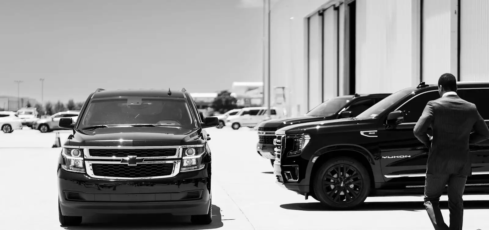 Man in a suit walking past a line of black SUVs, in black and white